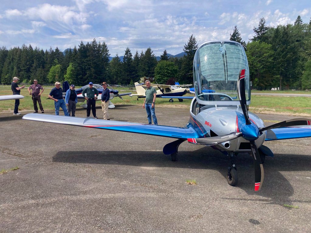 A blue and white low-wing airplane on the ramp at Packwood, with members talking in the background