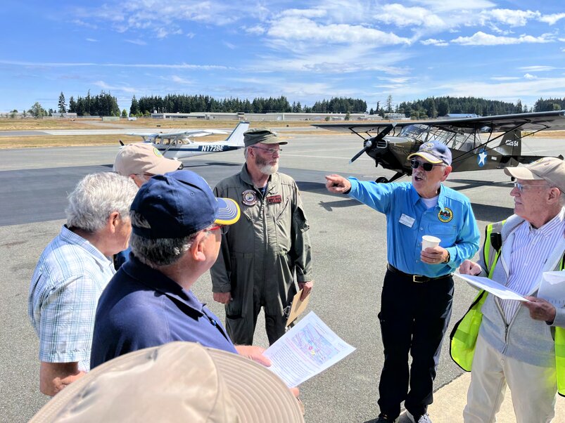 A volunteer in a blue shirt points across the ramp as he briefs pilots holding maps, with a Cessna and an olive drab Piper Cub parked behind them