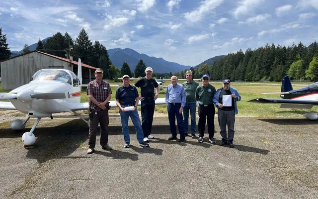 Smiling fly-out attendees lined up in front of a white low-wing airplane at Packwood, with forested mountains behind